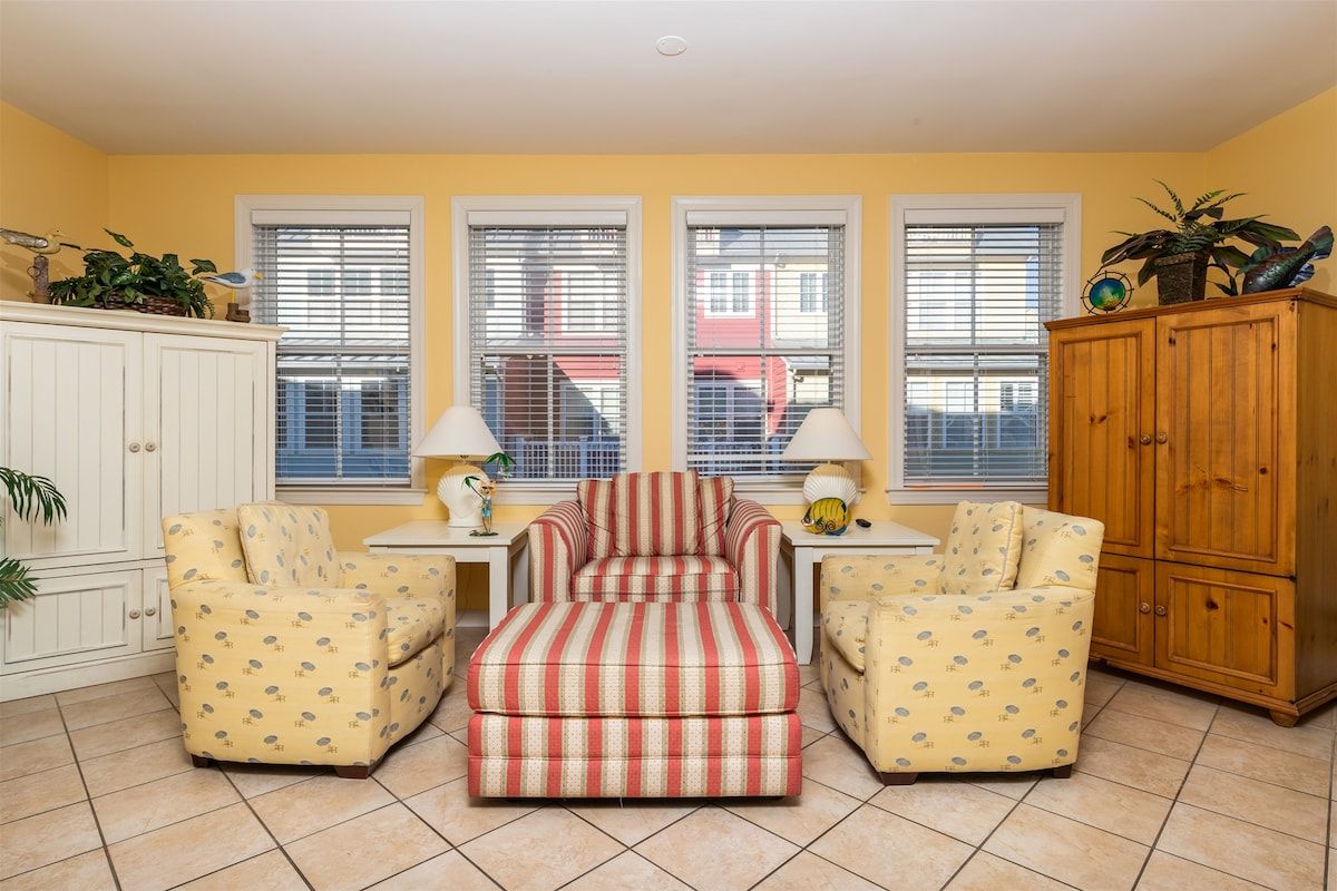 A living room with a striped ottoman, two patterned armchairs, a wooden cabinet, and a window with blinds.