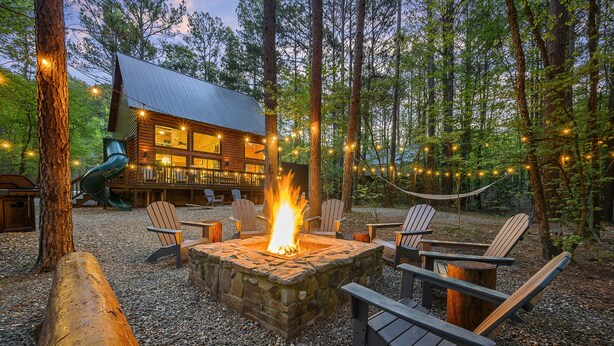 A cabin with a stone fire pit surrounded by Adirondack chairs, set in a forest with string lights. - Secluded Cabin: Sleeps 10, New Hot Tub, Fire Pit, Arcade, Foosball & Deck Slide (Broken Bow)