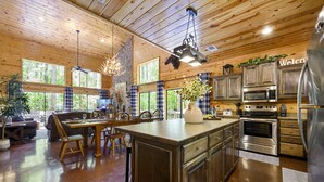 A spacious kitchen with wooden cabinets, stainless steel appliances, and large windows offering views of the forest. - Secluded Cabin: Sleeps 10, New Hot Tub, Fire Pit, Arcade, Foosball & Deck Slide (Broken Bow)