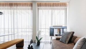 Ein modernes Wohnzimmer mit einer Couch, einem Couchtisch und einer Bank. Es gibt ein großes Fenster mit durchsichtigen Vorhängen, einen Fernseher auf einem Schrank und eine Pflanze auf dem Couchtisch.