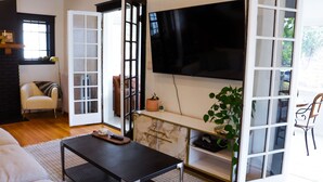 Uma sala de estar com sofá, mesa de centro, TV fixada na parede, armário com uma planta e porta que dá para outro cômodo.