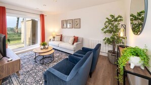 Ett vardagsrum med en soffa, en blå fåtölj, ett soffbord och en tv. Det finns ett fönster med gardiner, ett element och inramade tavlor på väggen.