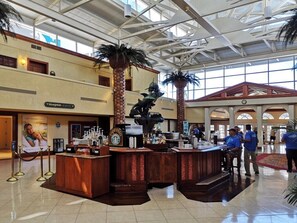 A spacious lobby with a central bar area, palm trees, and a high ceiling with skylights.