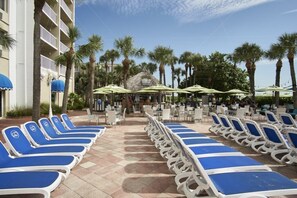 A poolside area with lounge chairs and umbrellas, surrounded by palm trees and a building with balconies.