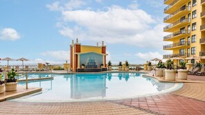 Una piscina con una fuente decorativa, rodeada de sillas de descanso y sombrillas, con un edificio al fondo.