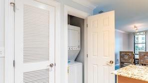 Un cuarto de lavado con lavadora y secadora, una puerta que da a un comedor con mesa y sillas, y vista a un edificio exterior.