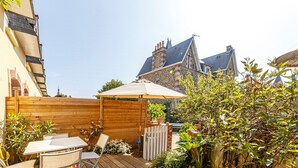A wooden deck with a table and chairs, surrounded by plants and a stone building in the background.