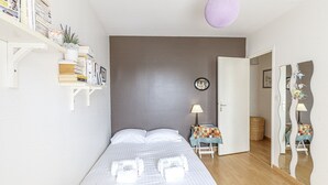 A bedroom with a bed, a bedside table with a lamp, a shelf with books and a small plant, and a mirror with a decorative frame.