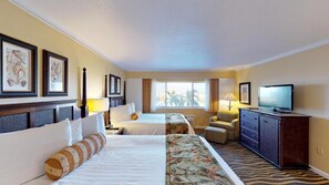 Two single beds with white linens and decorative pillows, a wooden dresser, and a television, set against a backdrop of framed artwork and a large window.