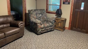 Een woonkamer met een bruine leren bank, een camouflage fauteuil, een klein houten kastje met een lamp en een deur met een bordje 'Welcome (Cabin)'.