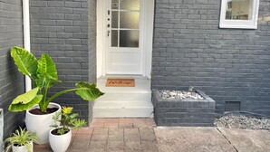 A dark grey brick house with a white front door, a small window, and a welcome mat. There are potted plants in front of the house. - In heart of mission Bay, Pet friendly, minutes walk to the beach (Auckland)