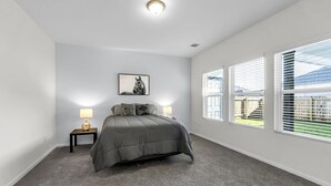 Ein Schlafzimmer mit einem Bett, zwei Nachttischlampen, einem gerahmten Bild an der Wand und großen Fenstern mit Jalousien.