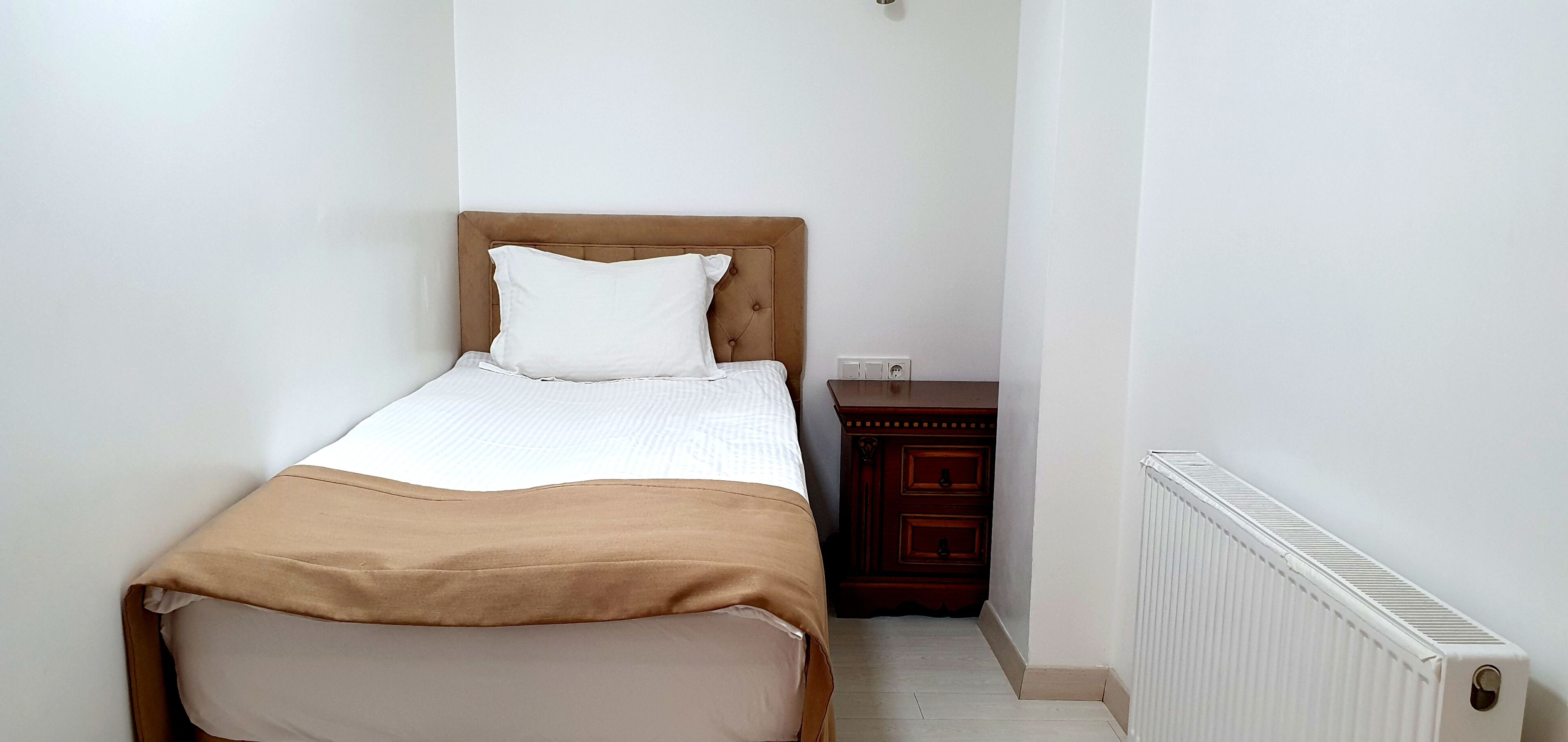 Una cama pequeña y ordenada con cabecera marrón y ropa de cama blanca en una habitación compacta, con un radiador y un pequeño armario de madera.