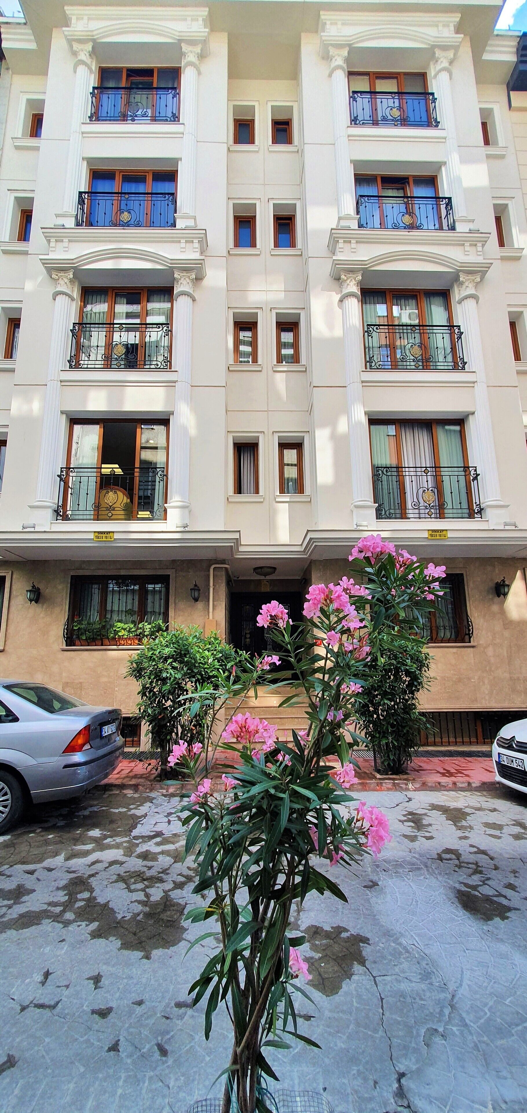 A multi-story building with balconies, a car parked in front, and pink flowers in the foreground.