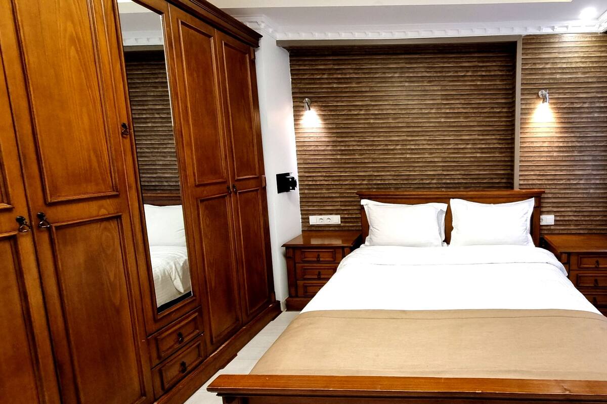 Une chambre à coucher avec un grand lit, une armoire en bois, deux tables de chevet et un luminaire fixé au mur.