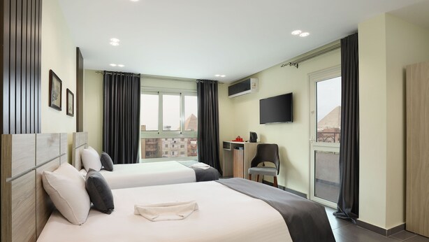 A hotel room with two beds, a TV, a desk with a chair, and a window with curtains. - Brothers Pyramids View (Giza)