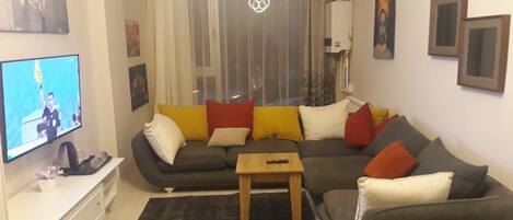 A living room with a sectional sofa, a coffee table, a flat-screen TV, and a window with curtains.