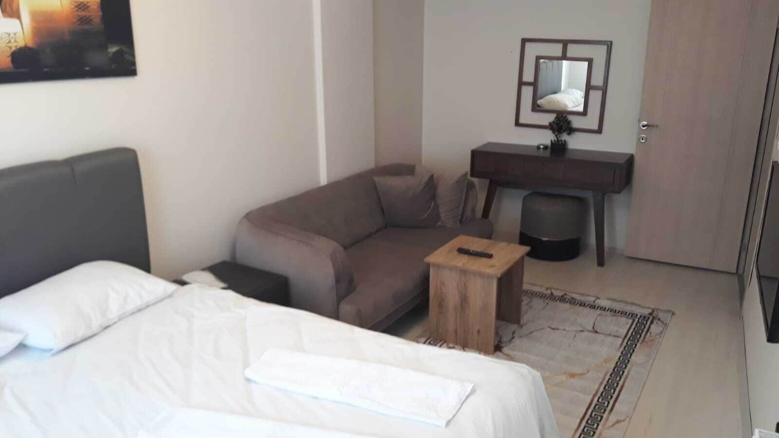 A hotel room with a bed, a sofa, a small wooden stool, a desk with a lamp, and a mirror on the wall.