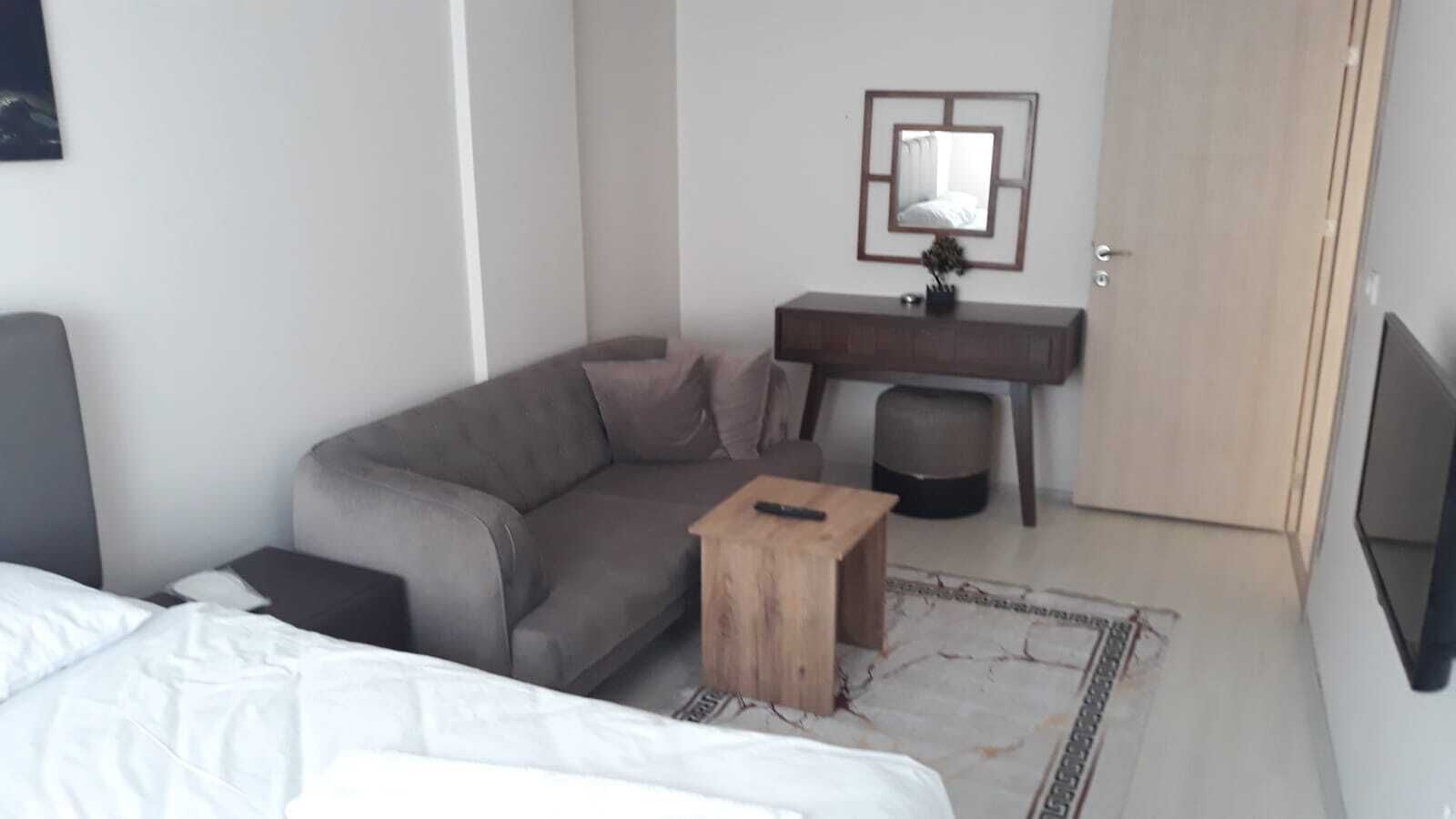 Een moderne hotelkamer met een grijze bank, een houten bijzettafeltje, een klein bureau met spiegel en een flatscreen televisie aan de muur.