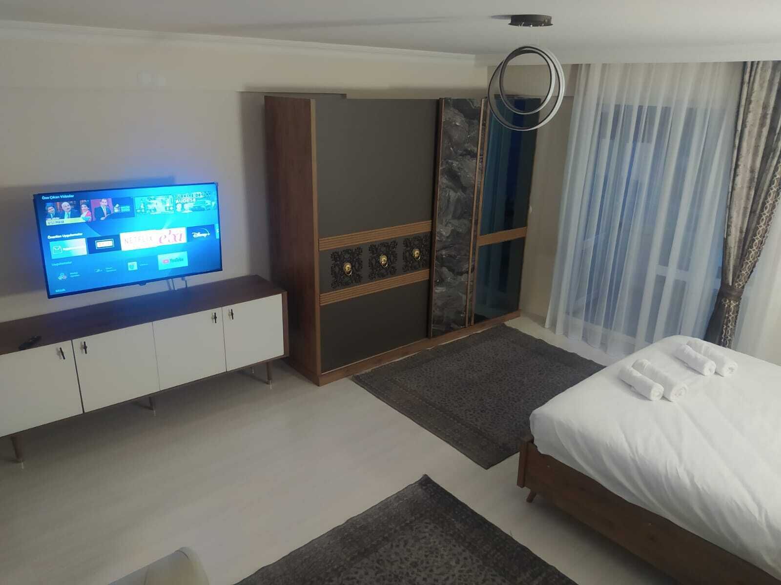 Una habitación de hotel moderna con una televisión de pantalla plana sobre un mueble blanco, una cama con ropa de cama blanca, un armario con un panel que imita el mármol y un amplio ventanal con cortinas translúcidas.