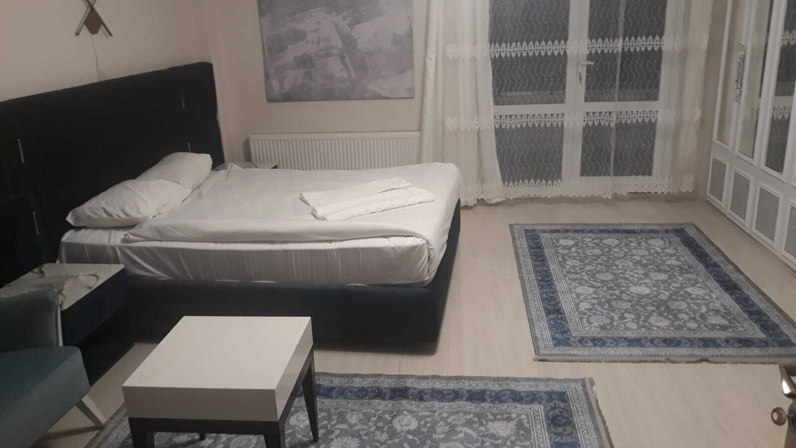 Een slaapkamer met een bed, nachtkastje, stoel en raam met kantgordijnen.