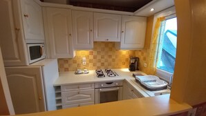 A compact kitchen with white cabinets, a microwave, a coffee maker, and a window with yellow curtains. - Apartment at the foot of DEAUVILLE/TROUVILLE (Touques)