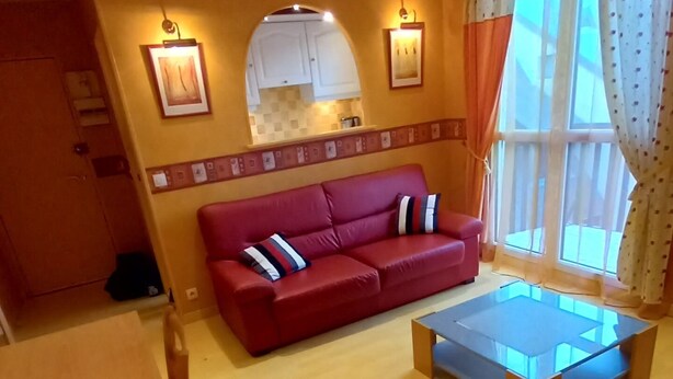 A living room with a red sofa, a glass coffee table, and a dining area with wooden furniture. - Apartment at the foot of DEAUVILLE/TROUVILLE (Touques)