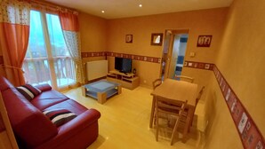 A living room with a red sofa, a TV, a small table, and a dining area with chairs. - Apartment at the foot of DEAUVILLE/TROUVILLE (Touques)