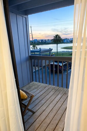 A balcony with a wooden floor and a chair, overlooking a parking area and a body of water with buildings in the background. - Ocean View Pet-Friendly Condo- 5-Min Walk to Boardwalk- Lost of Summer available (Carolina Beach)