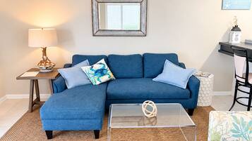 Ein Wohnzimmer mit einem blauen Sofa, einem Glastisch, einer Lampe und einem gerahmten Bild an der Wand.