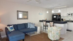 A living room with a blue sofa, a glass coffee table, a floral armchair, and a kitchen area with a black refrigerator, microwave, and bar stools.