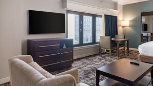 A hotel room with a bed, a desk, a chair, a dresser, a TV, and a window with a city view.