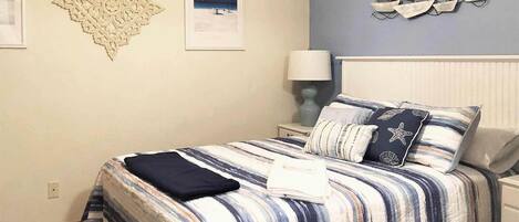 A neatly made bed with striped bedding and starfish pillows, a wall-mounted ship decoration, and a framed picture of a beach scene.