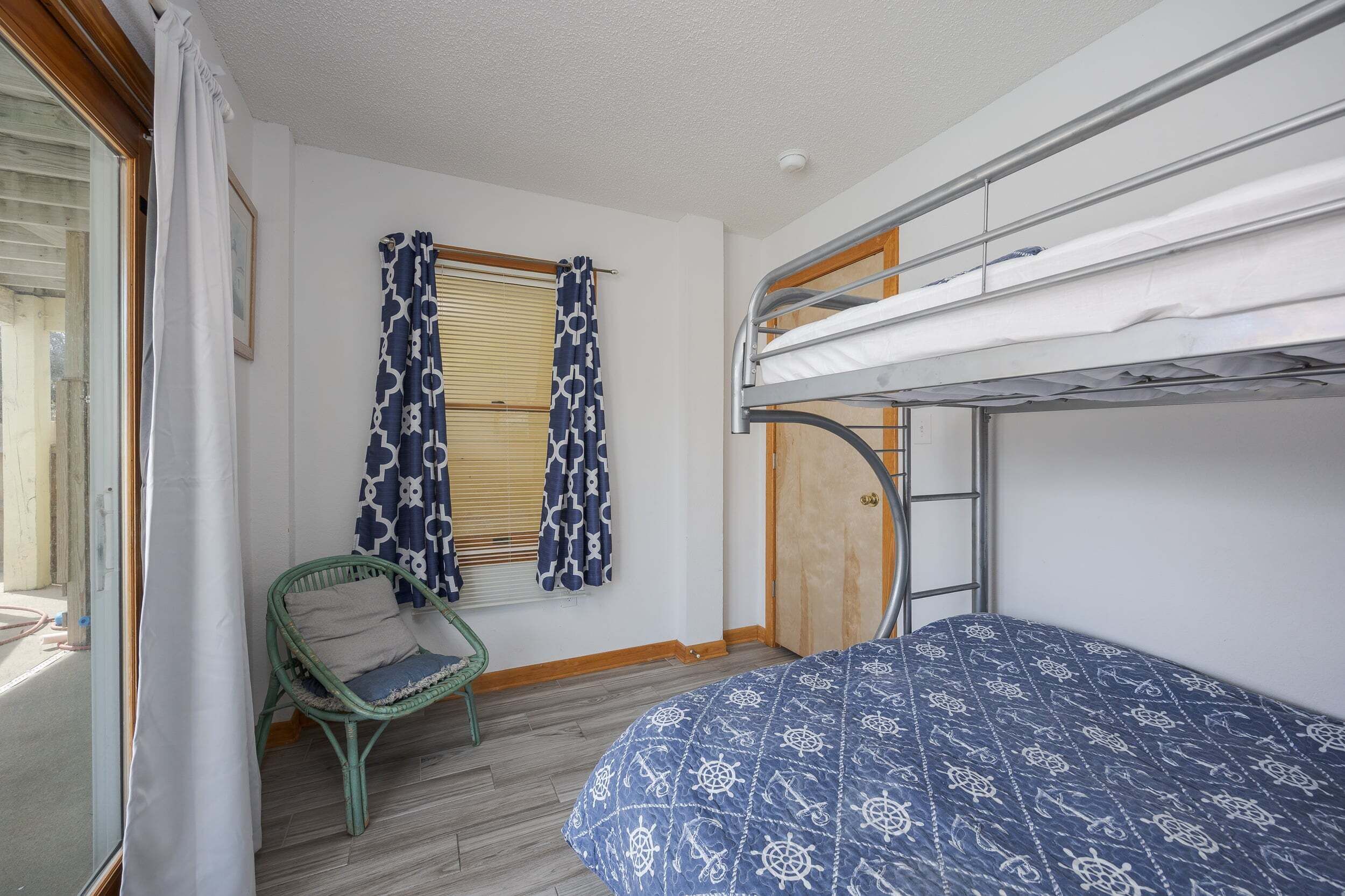 Habitación con literas, una colcha estampada, una silla de mimbre, una ventana con cortinas decoradas y una puerta.