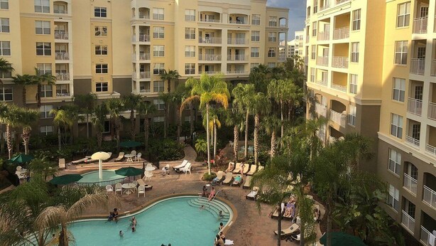 A hotel with multiple balconies, a central swimming pool, and surrounding palm trees.