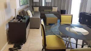 A modern living room with a sofa set, a glass coffee table, and a dining table with chairs. There are two lamps, a TV, and a cabinet.