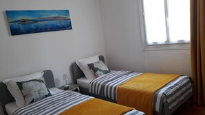 A room with two beds, each with striped bedding and a yellow throw blanket. There is a painting on the wall depicting a coastal scene. - Cozy villa in the heart of the Luberon (Apt)
