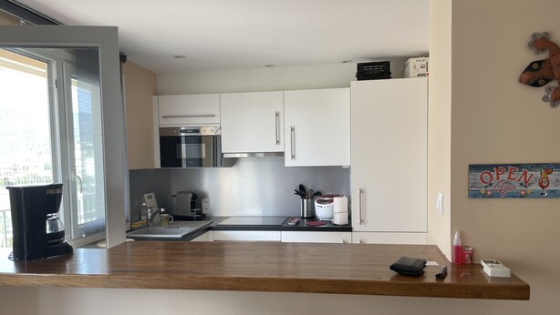 A modern kitchen with white cabinets, a built-in oven, and a coffee machine. There is a wooden countertop and a dining area with two chairs.