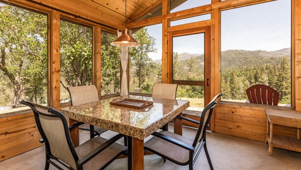 A wooden interior with a dining table, chairs, and a view of a forested landscape through large windows.