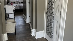 Un couloir au plancher de bois mène à une chambre à coucher où se trouvent un lit et une commode. Une porte est ornée d’un rideau à motifs floraux, et un fer à repasser blanc est posé sur un porte-fer fixé au mur.