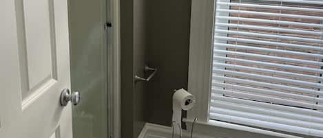 A bathroom with a shower, toilet, and window with blinds.