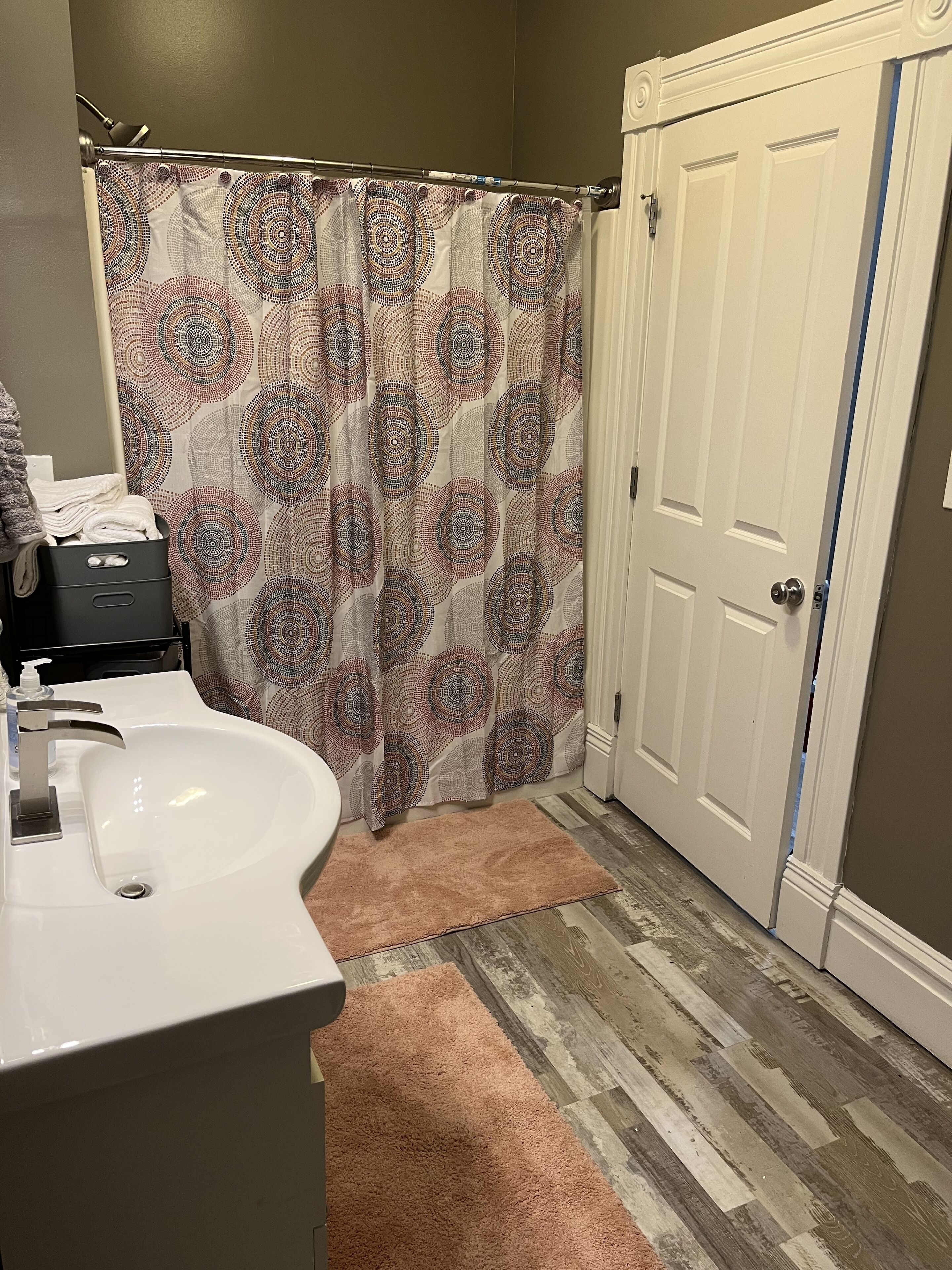 A bathroom with a white sink, a patterned shower curtain, a wooden floor, and a door with a blue stripe.
