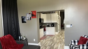 Un salon moderne avec un canapé gris, un fauteuil noir et un plaid rouge. On aperçoit, au fond, une cuisine avec des meubles blancs et un four à micro-ondes.