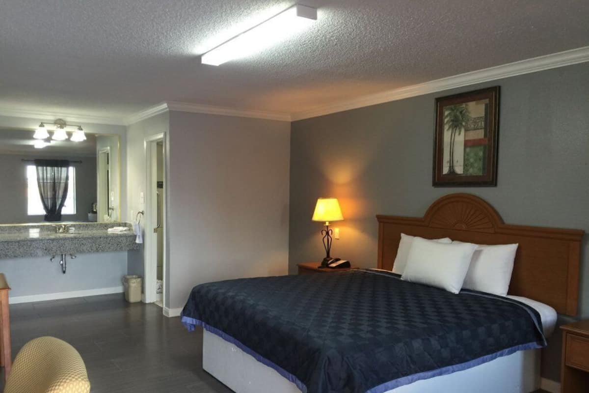 A hotel room with a bed, a nightstand, a lamp, a bathroom with a sink and mirror, and a painting on the wall.
