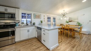Een moderne keuken met witte kasten, een ingebouwde magnetron en een vaatwasser. Er is een eethoek met een houten tafel en stoelen, en een kroonluchter erboven.