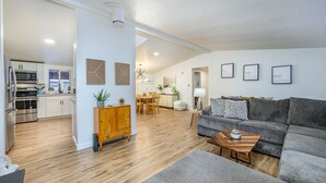 Een moderne woonkamer met een grijze bank, een houten salontafel en een eethoek met een houten tafel en stoelen. Er is een keuken met witte kastjes en roestvrijstalen apparatuur.