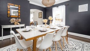Un comedor con una mesa blanca dispuesta para cuatro personas, rodeada de sillas blancas. Un espejo decorativo y una lámpara de tejido están en las paredes.