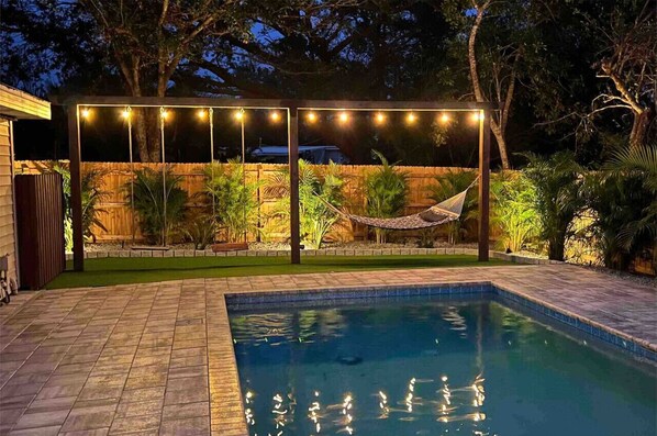 Relax by the clear pool surrounded by a deck, hammock, and lush tropical plants.