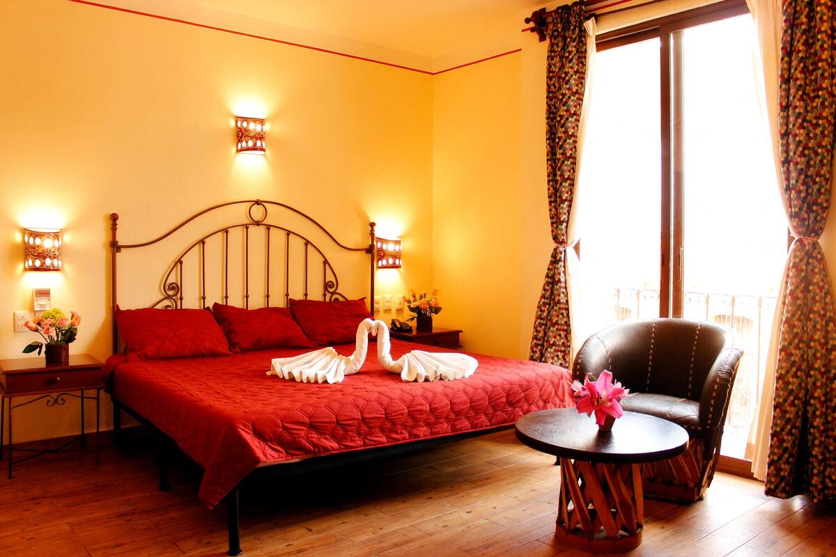 Una habitación de hotel con una cama grande, dos mesitas de noche, una silla y un balcón con cortinas.