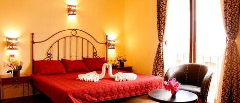 Quarto de hotel com cama grande, duas mesas de cabeceira, uma cadeira e varanda com cortinas.